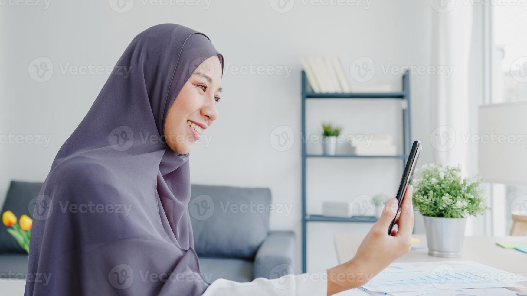 Young Asia muslim businesswoman using smart phone talk to friend by videochat brainstorm online meeting while remotely work from home at living room. Social distancing, quarantine for corona virus. photo