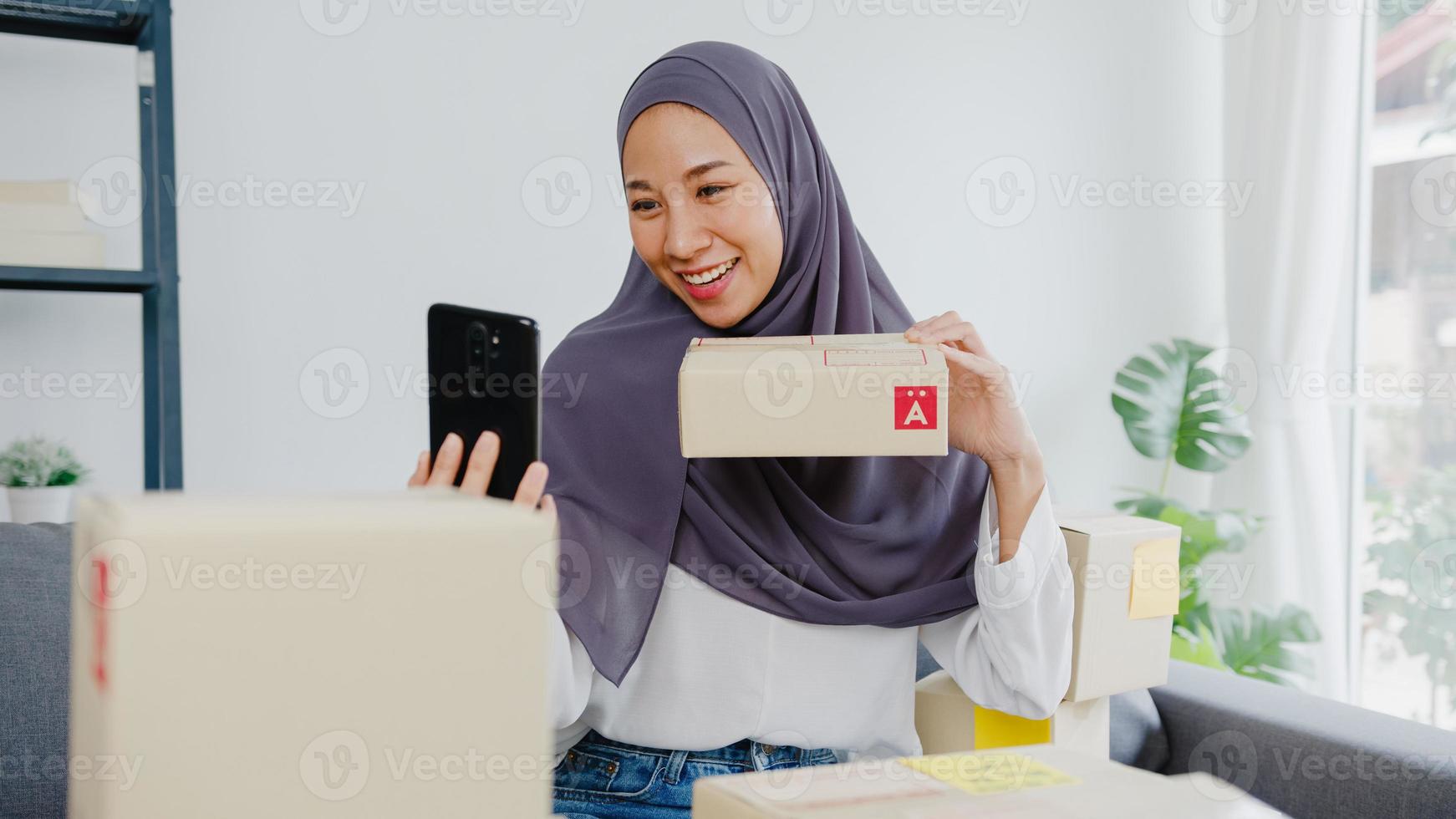 Young Asia muslim businesswoman blogger using mobile phone camera to recording vlog video live streaming review product at home office. Small business owner, start up online market delivery concept. photo