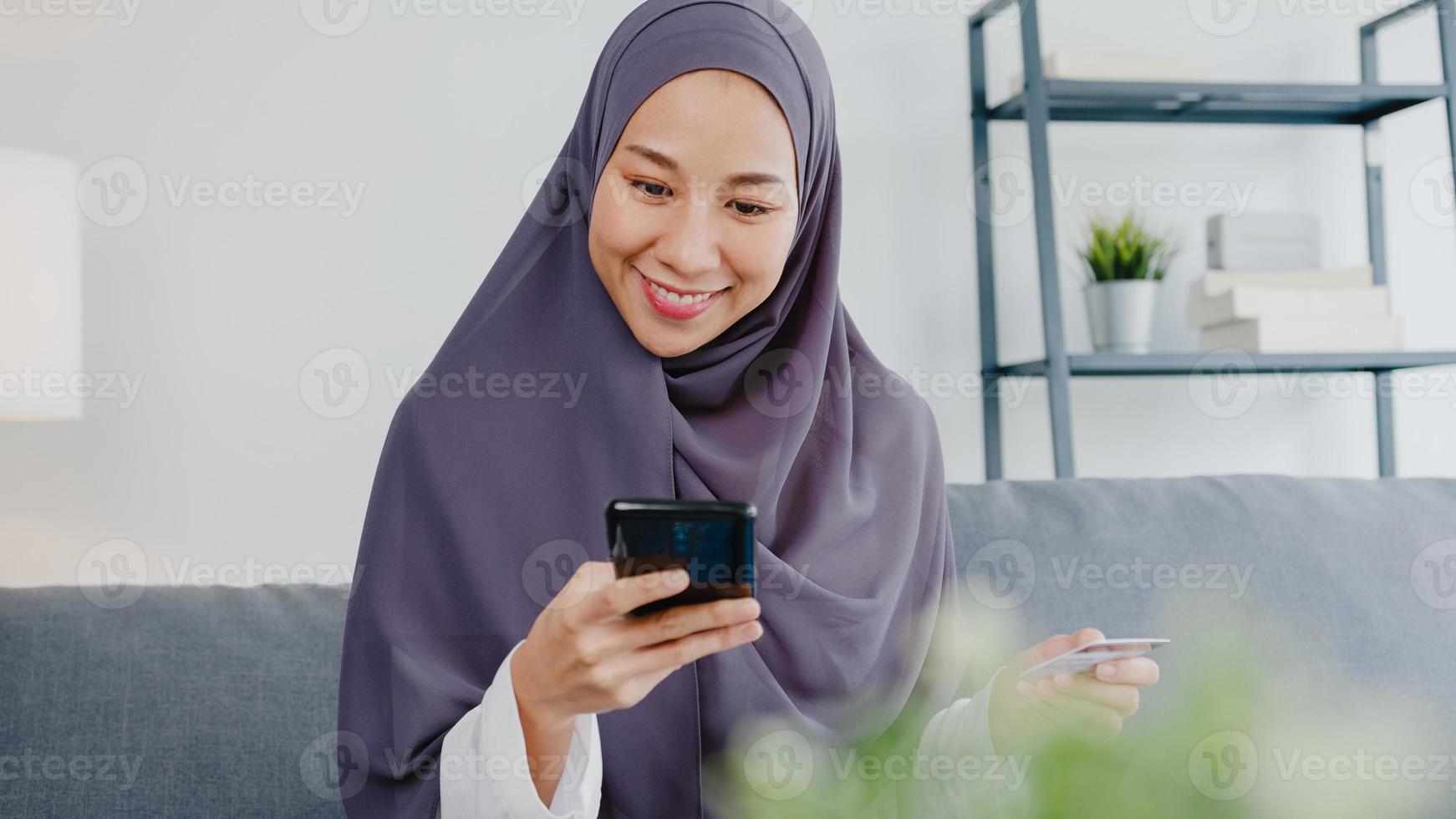 Asia muslim lady use smart phone, credit card buy and purchase e-commerce internet in living room at house. Stay at home, online shopping, self isolation, social distance, quarantine for coronavirus. photo