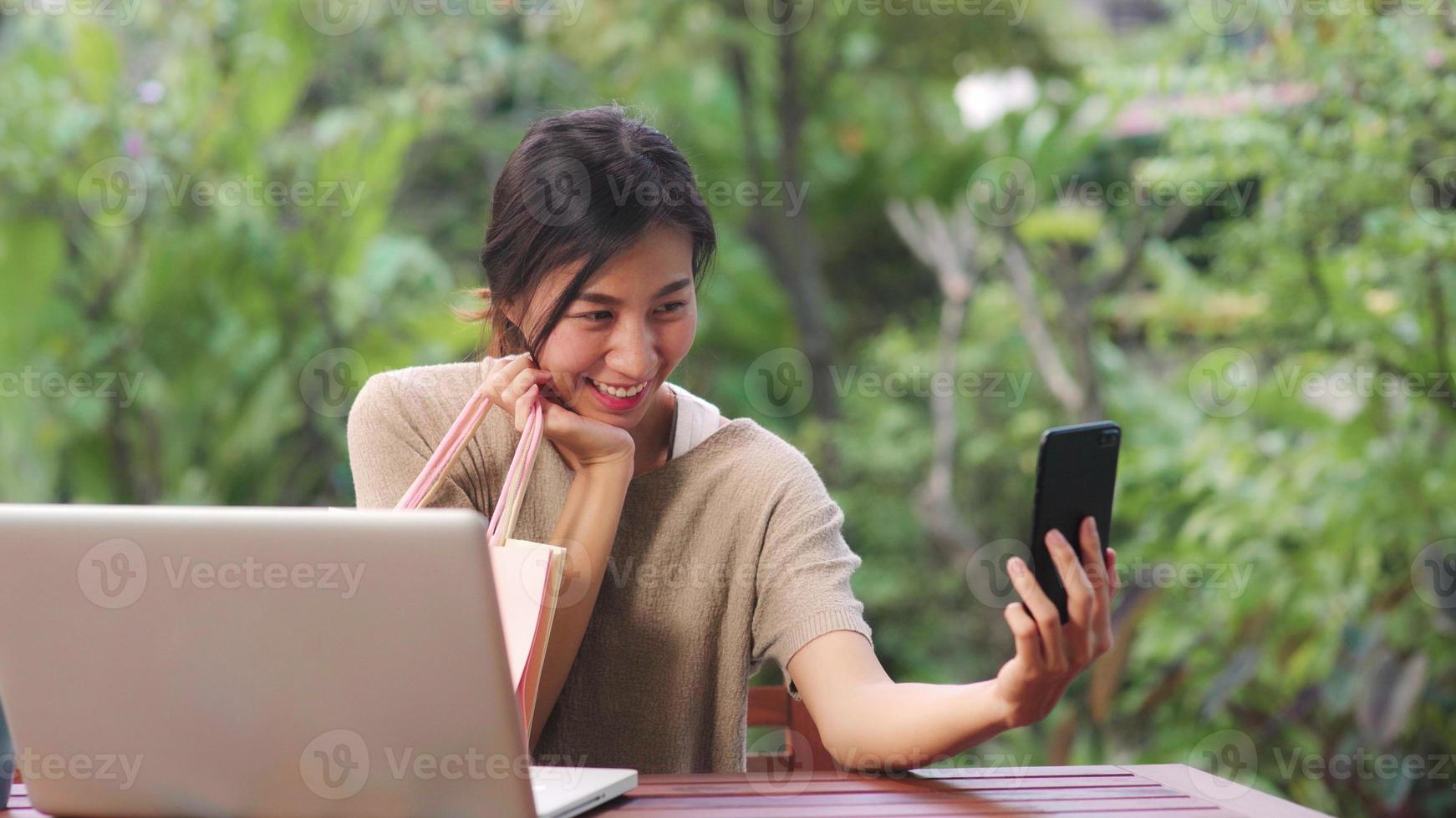 Asian woman using mobile phone selfie post in social media, female relax feeling happy showing shopping bags sitting on table in the garden in morning. Lifestyle women relax at home concept. photo