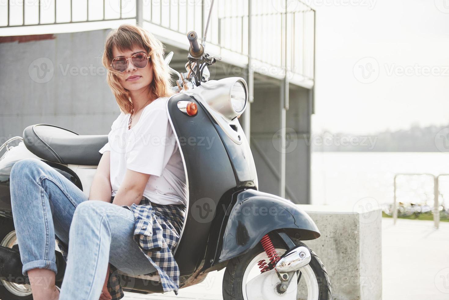 Retrato de una hermosa chica hipster sentada en un scooter retro negro, sonriendo posando y disfrutando del cálido sol primaveral. foto