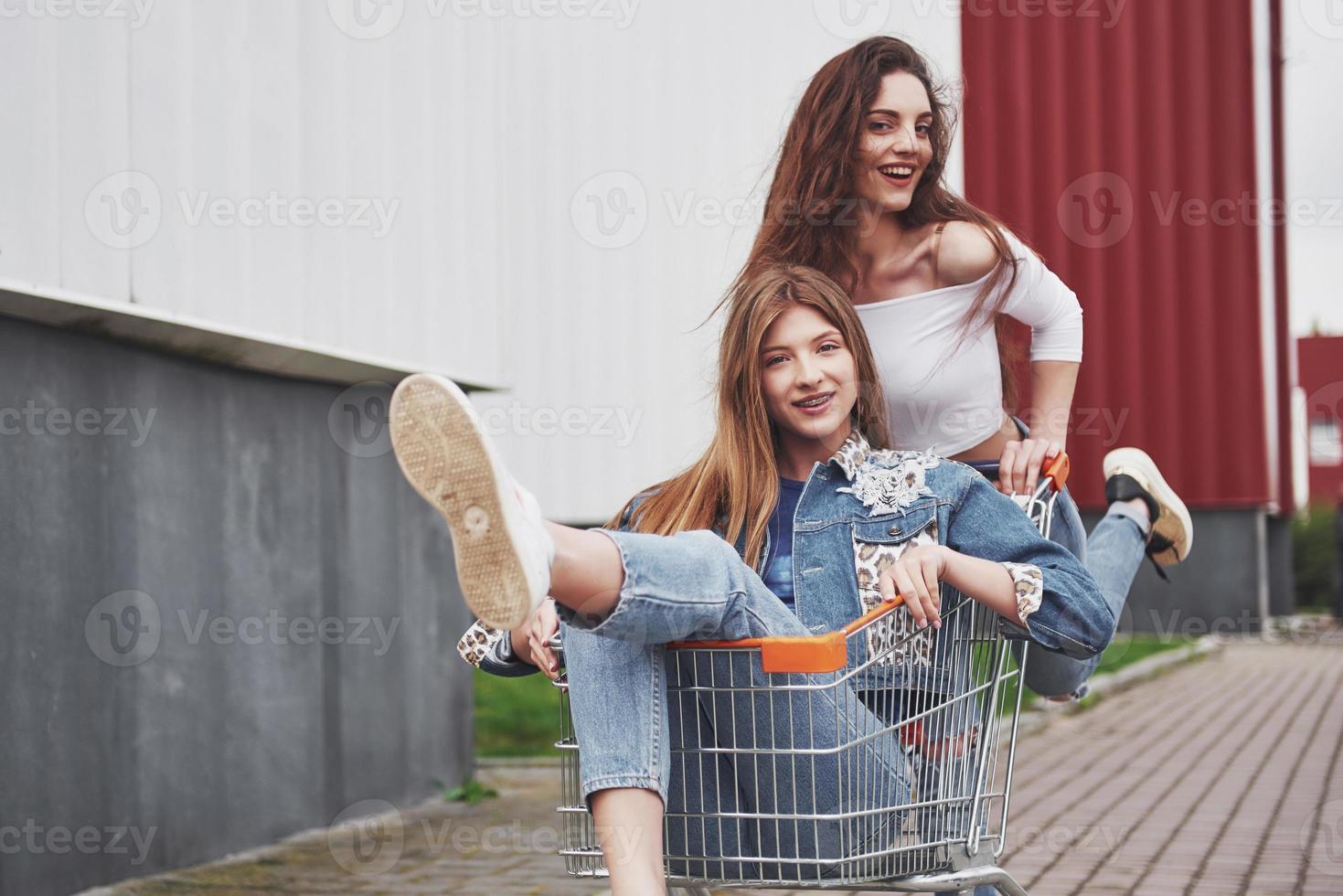 Two young happy women having fun shopping trolley race outdoors. photo