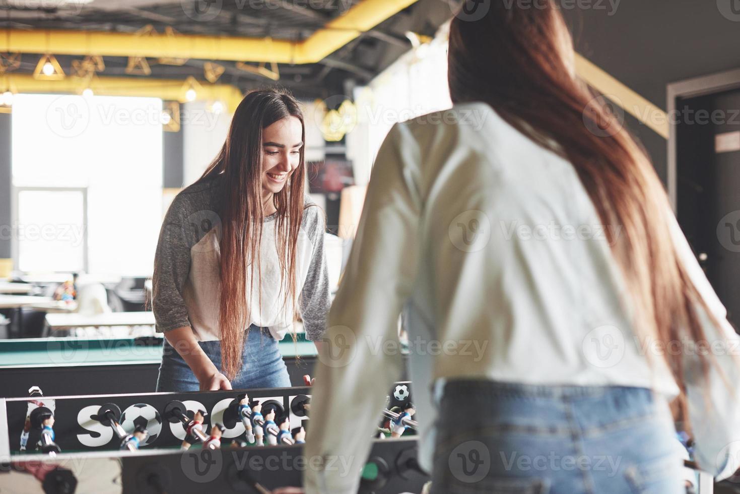 dos hermosas niñas gemelas juegan al futbolín y se divierten. una de las hermanas sostiene una pelota de juguete en su mano y muestra la lengua foto