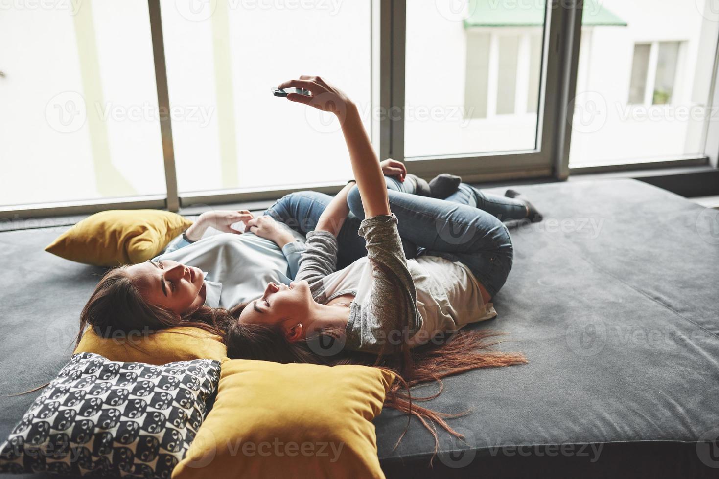 dos lindas hermanas gemelas sonrientes sosteniendo smartphone y haciendo selfie. las niñas se acuestan en el sofá posando y alegría foto