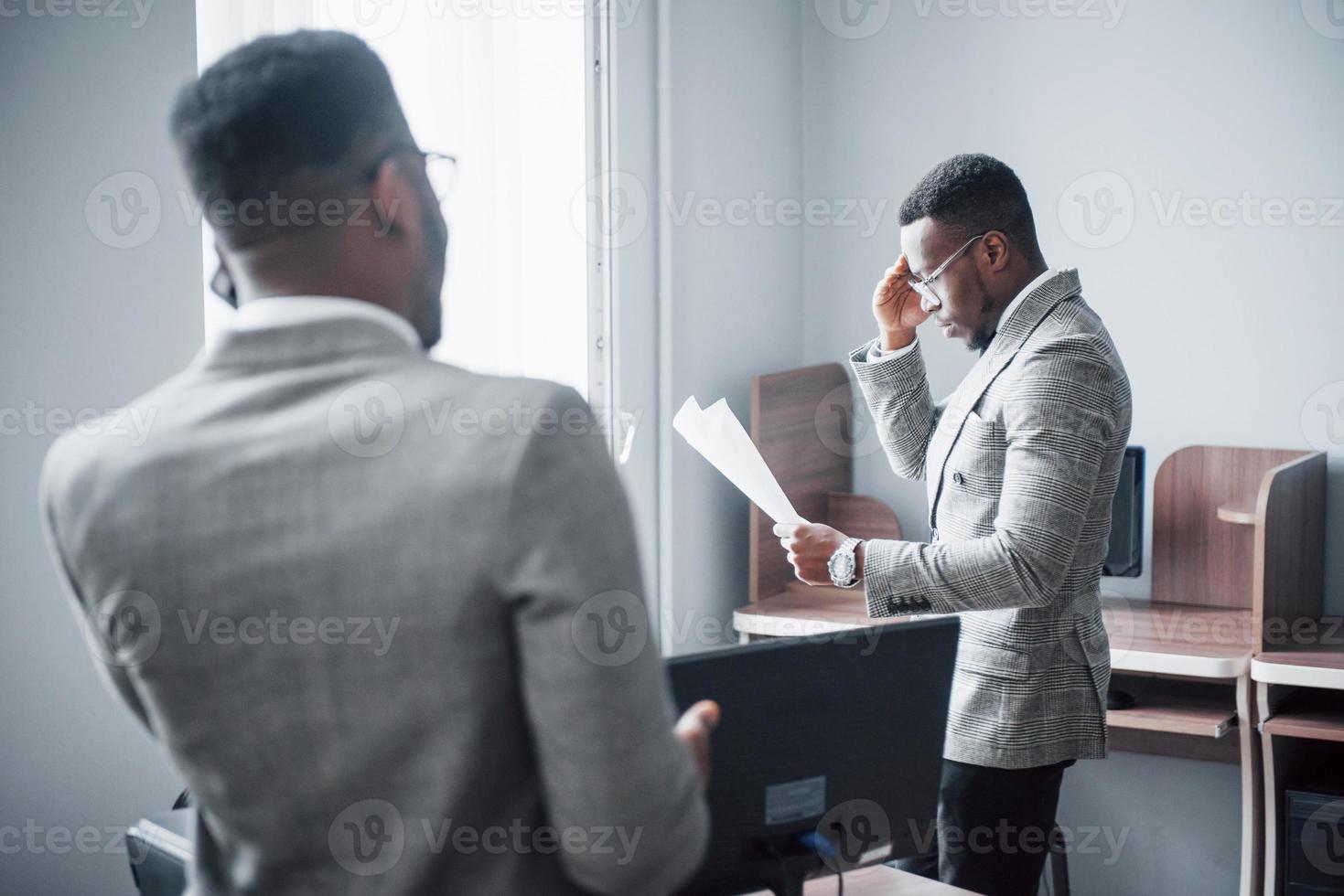 empresario moderno en el trabajo. Dos hombres de negocios confiados en ropa formal discutiendo algo mientras uno de ellos mira los documentos y el segundo una importante llamada telefónica. foto