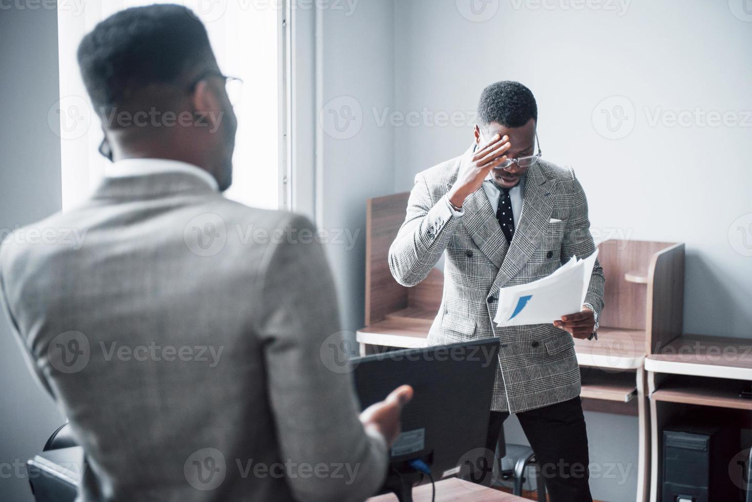 empresario moderno en el trabajo. Dos hombres de negocios confiados en ropa formal discutiendo algo mientras uno de ellos mira los documentos y el segundo una importante llamada telefónica. foto