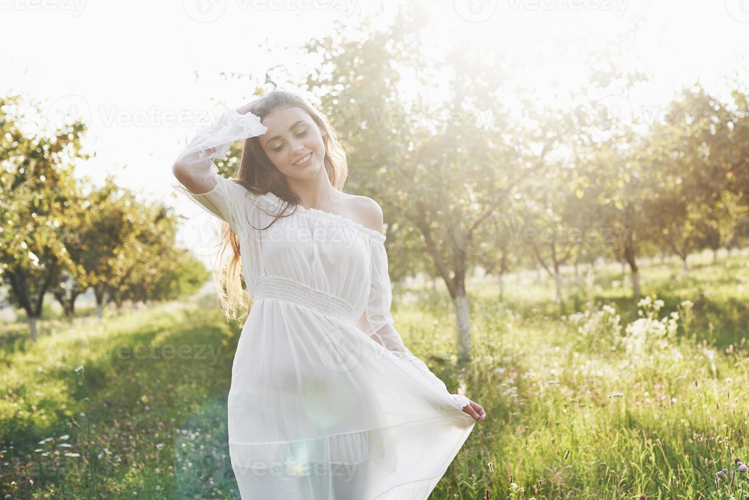 una niña con un vestido largo blanco está caminando en el jardín. hermosa puesta de sol a través de las hojas de los árboles foto