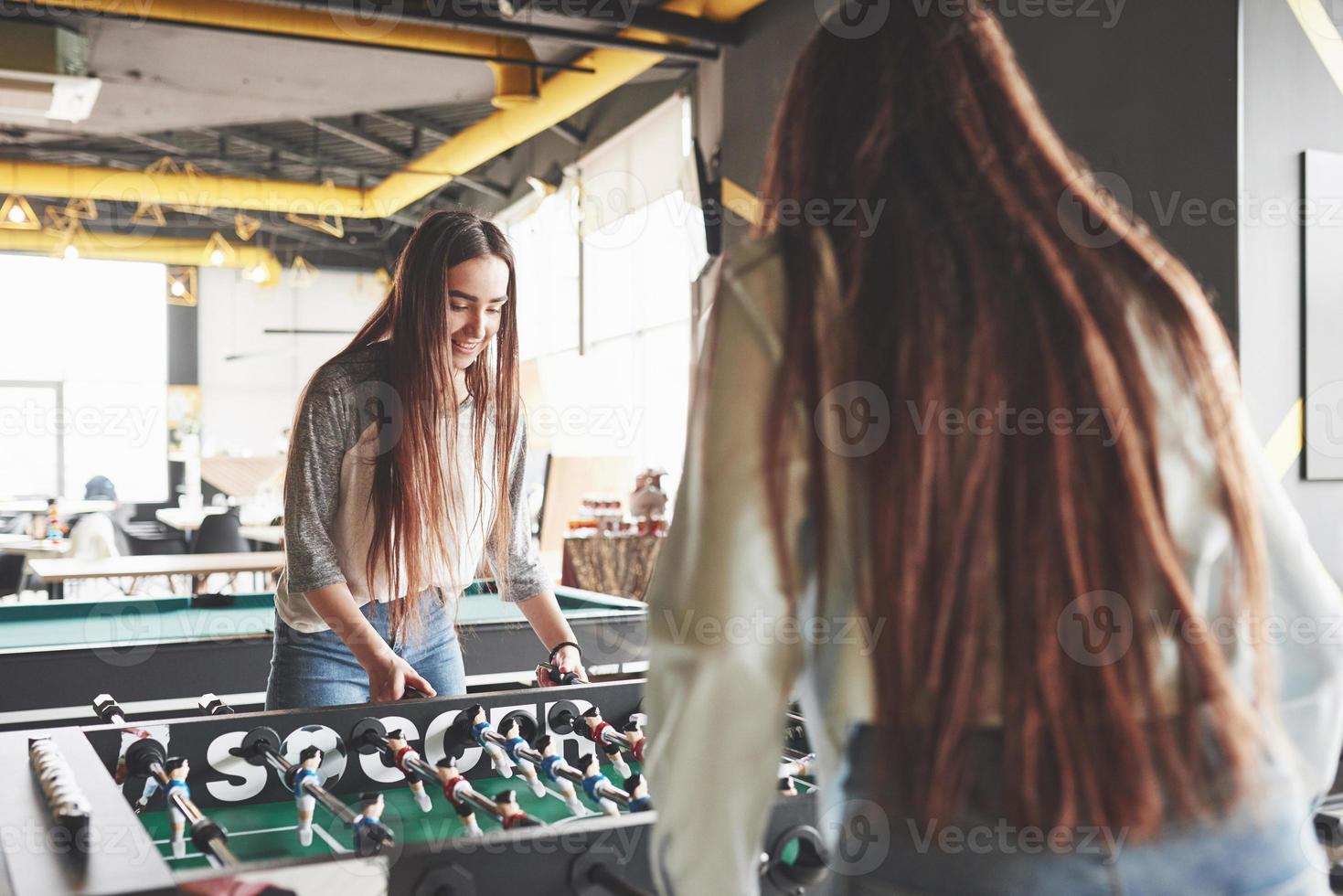 dos hermosas niñas gemelas juegan al futbolín y se divierten. una de las hermanas sostiene una pelota de juguete en su mano y muestra la lengua foto