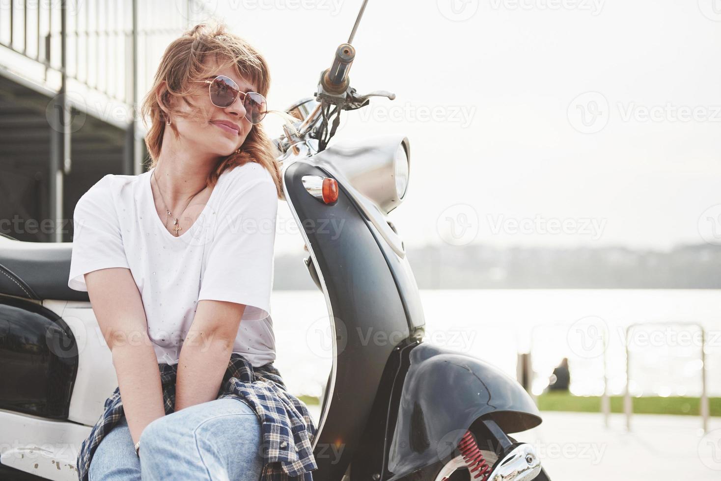 Retrato de una hermosa chica hipster sentada en un scooter retro negro, sonriendo posando y disfrutando del cálido sol primaveral. foto