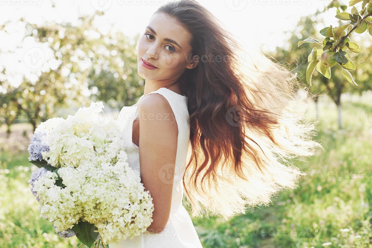 una hermosa joven con un vestido blanco claro y un ramo de flores de verano pone un buen día en el jardín foto