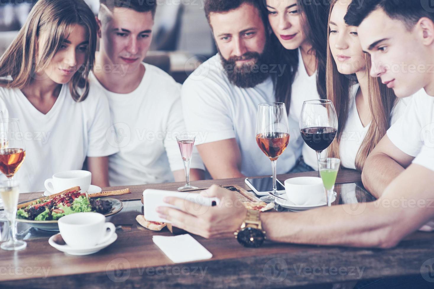 A group of people make a selfie photo in a cafe. The best friends gathered together at a dinner table eating pizza and singing various drinks