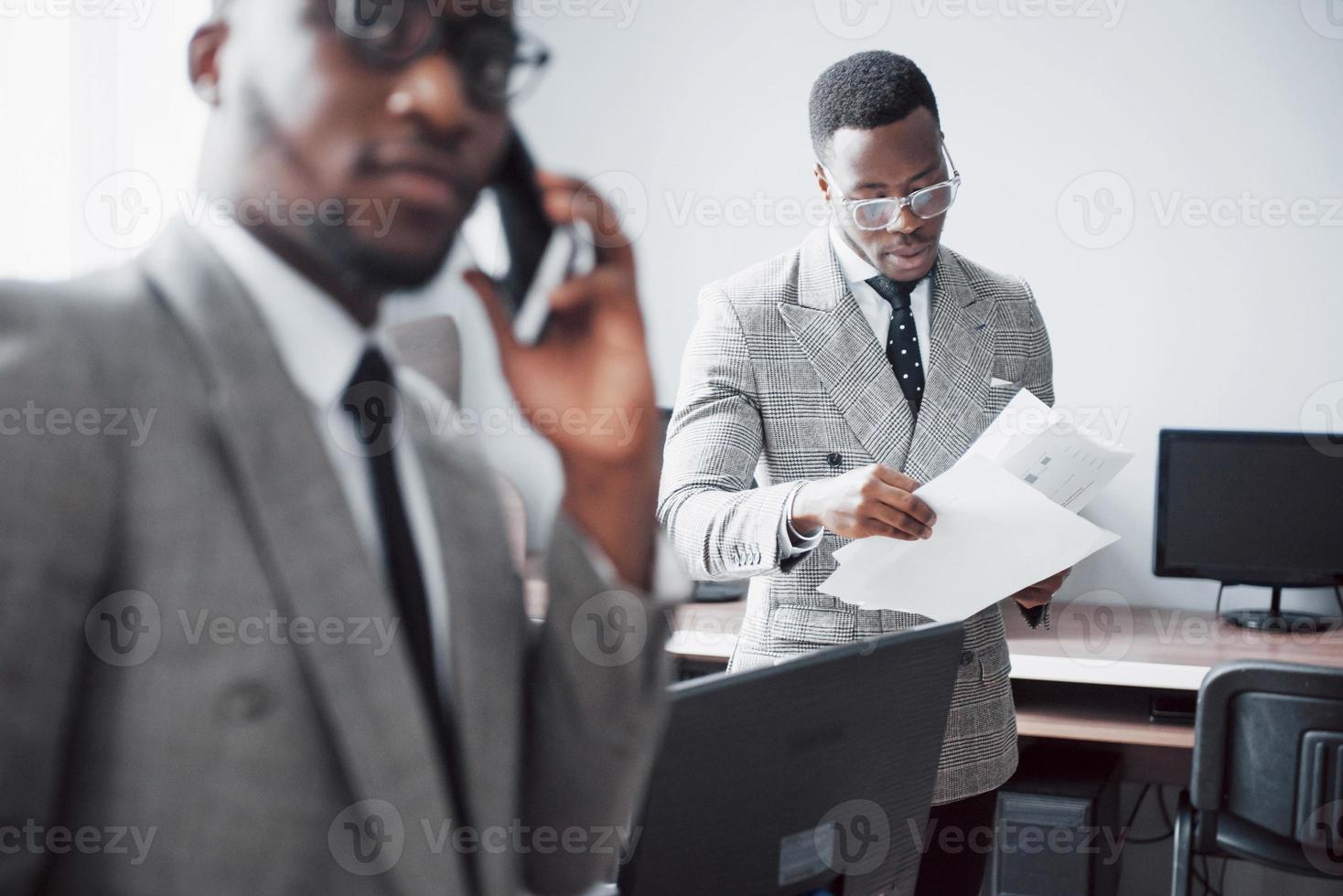 empresario moderno en el trabajo. Dos hombres de negocios confiados en ropa formal discutiendo algo mientras uno de ellos mira los documentos y el segundo una importante llamada telefónica. foto