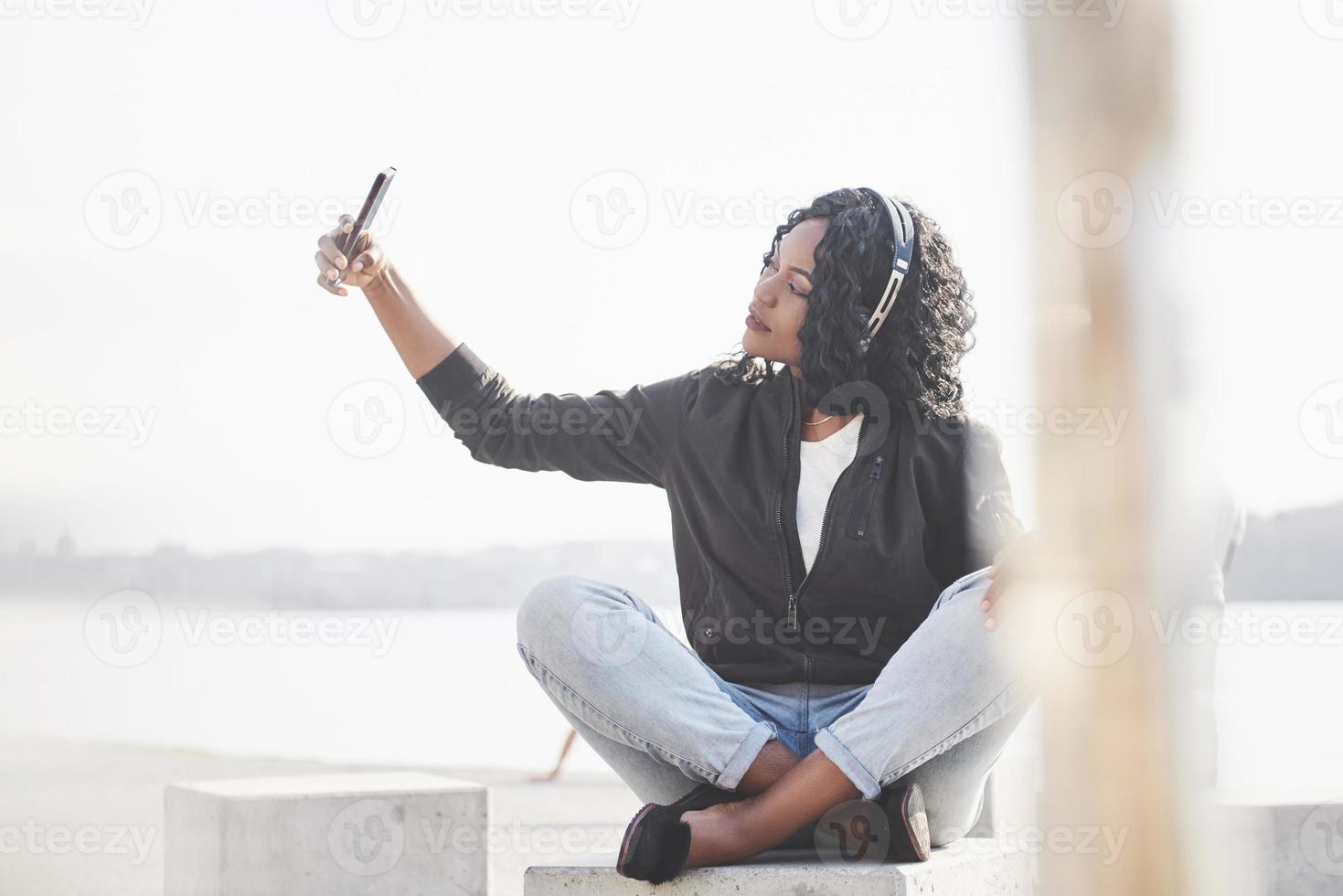 Portrait of a beautiful young pretty African American girl sitting on the beach or lake and listening to music in her headphones photo