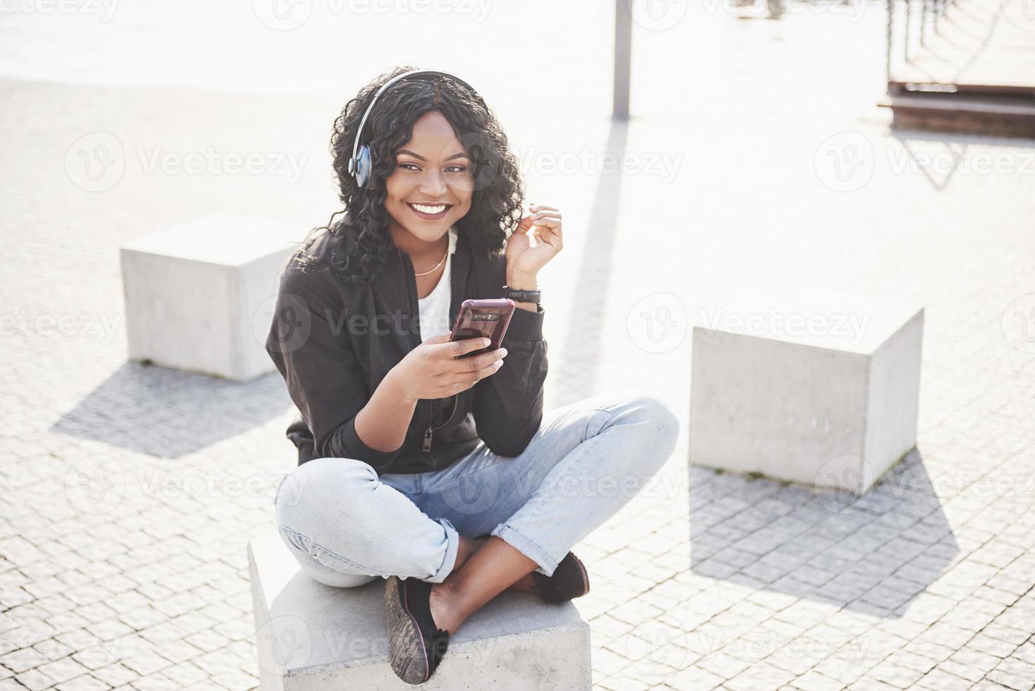 Portrait of a beautiful young pretty African American girl sitting on the beach or lake and listening to music in her headphones photo