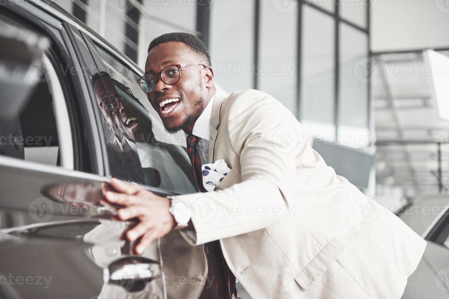 apuesto hombre negro en el concesionario está abrazando su nuevo coche y sonriendo foto