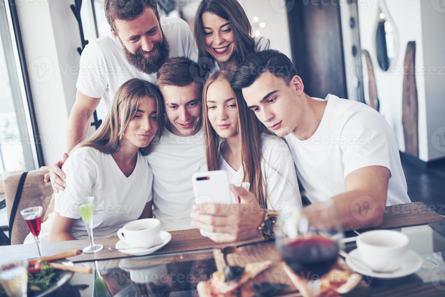 A group of people make a selfie photo in a cafe. The best friends gathered together at a dinner table eating pizza and singing various drinks