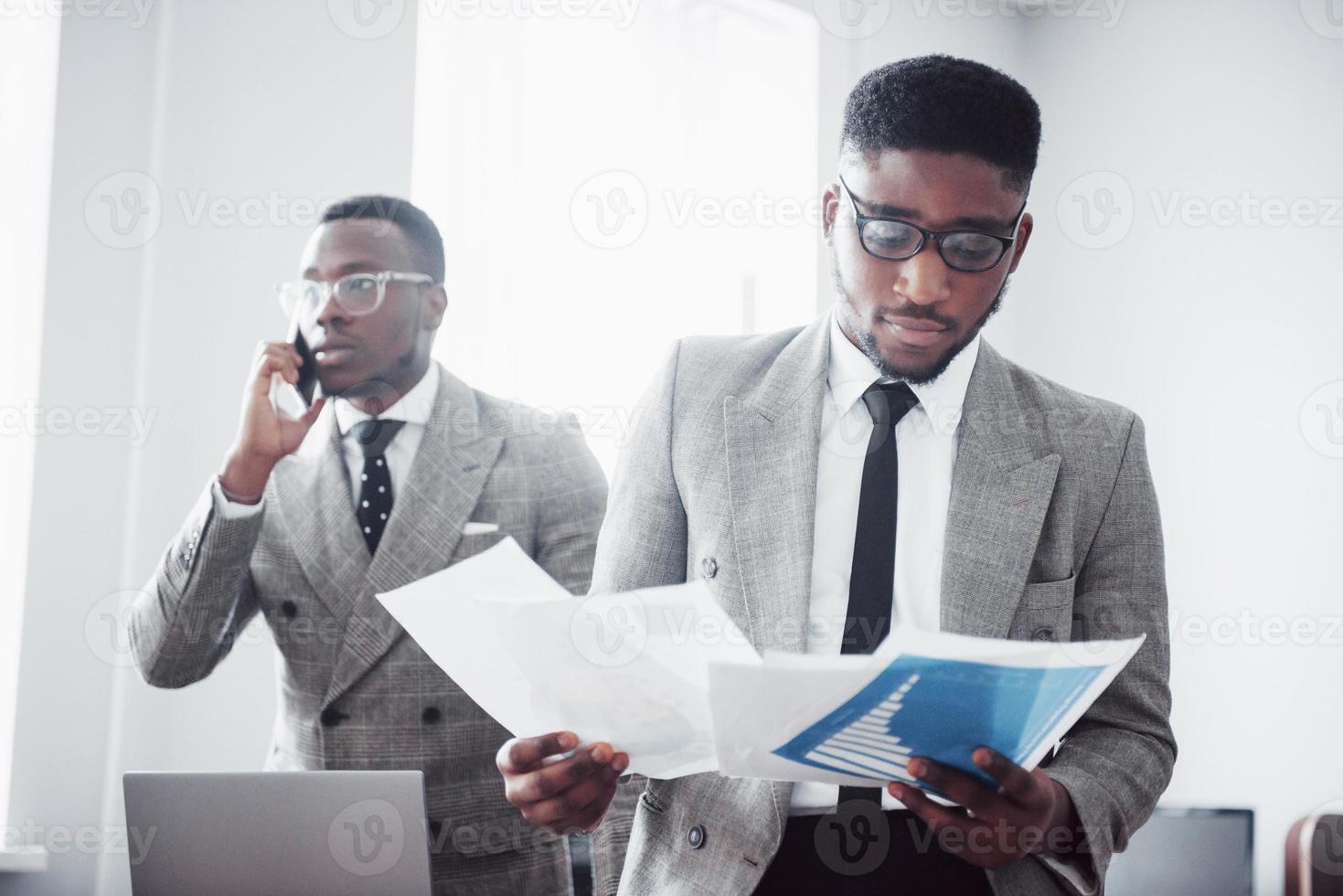 empresario moderno en el trabajo. Dos hombres de negocios confiados en ropa formal discutiendo algo mientras uno de ellos mira los documentos y el segundo una importante llamada telefónica. foto