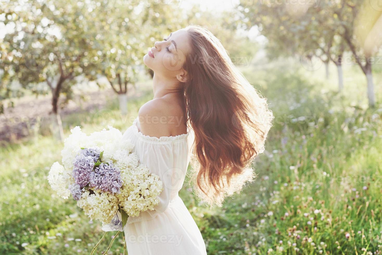 una hermosa joven con un vestido blanco claro y un ramo de flores de verano pone un buen día en el jardín foto