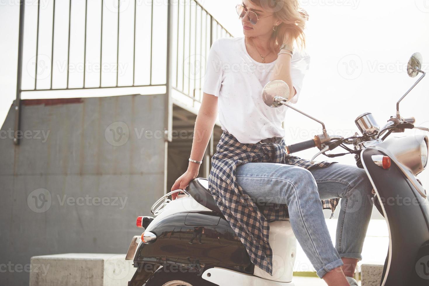 Retrato de una hermosa chica hipster sentada en un scooter retro negro, sonriendo posando y disfrutando del cálido sol primaveral. foto