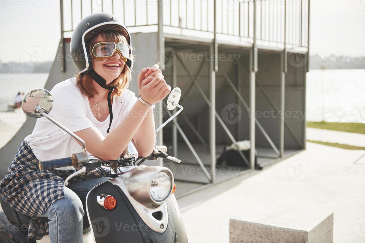 Retrato de una hermosa chica hipster sentada en un scooter retro negro, sonriendo posando y disfrutando del cálido sol primaveral. foto