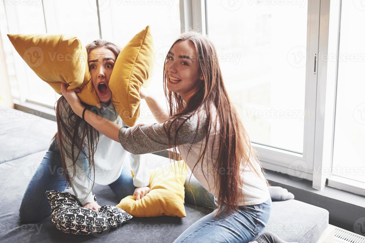 Two beautiful young twins sisters spending time together and are battling with pillows. Siblings having fun at home concept photo