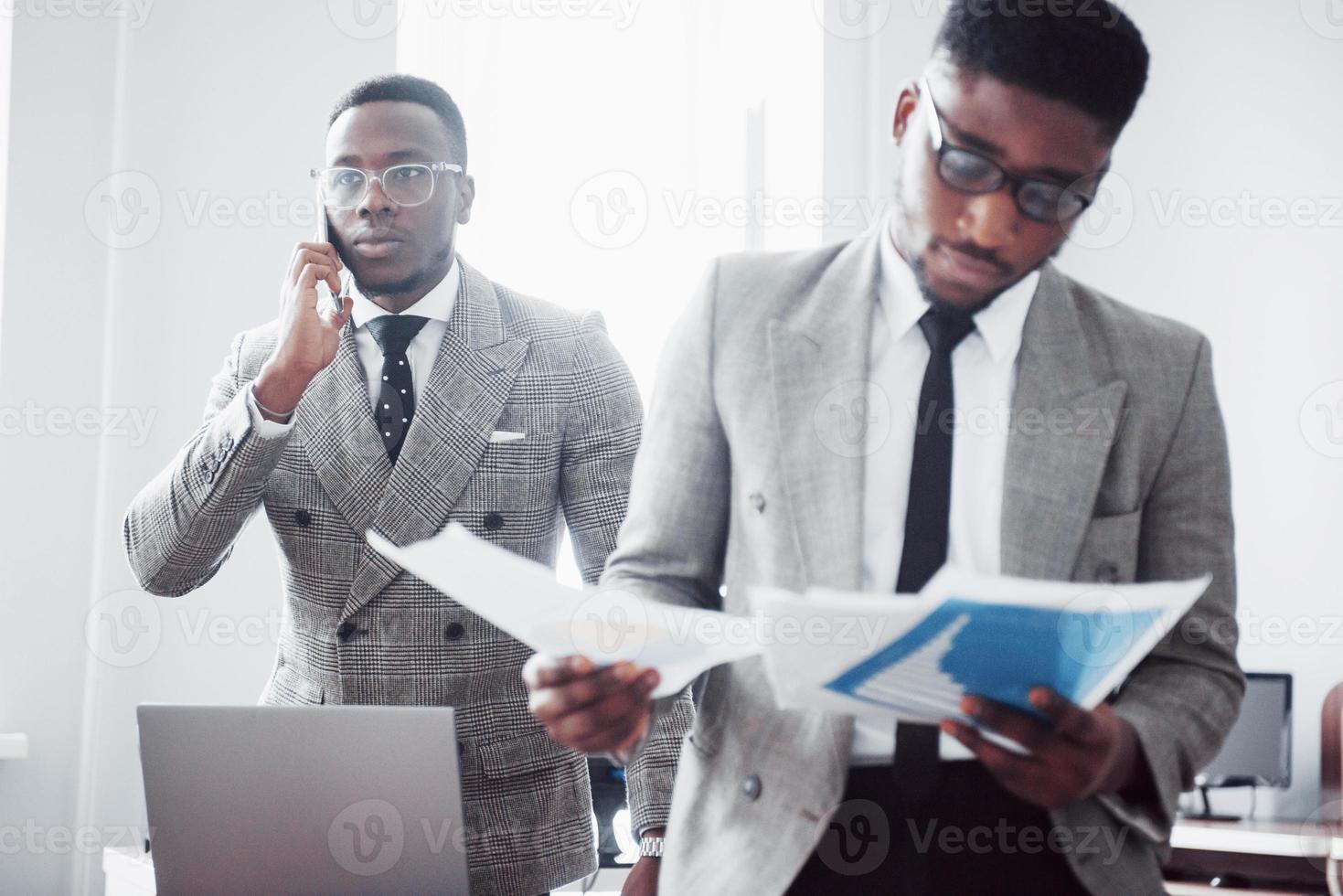 empresario moderno en el trabajo. Dos hombres de negocios confiados en ropa formal discutiendo algo mientras uno de ellos mira los documentos y el segundo una importante llamada telefónica. foto