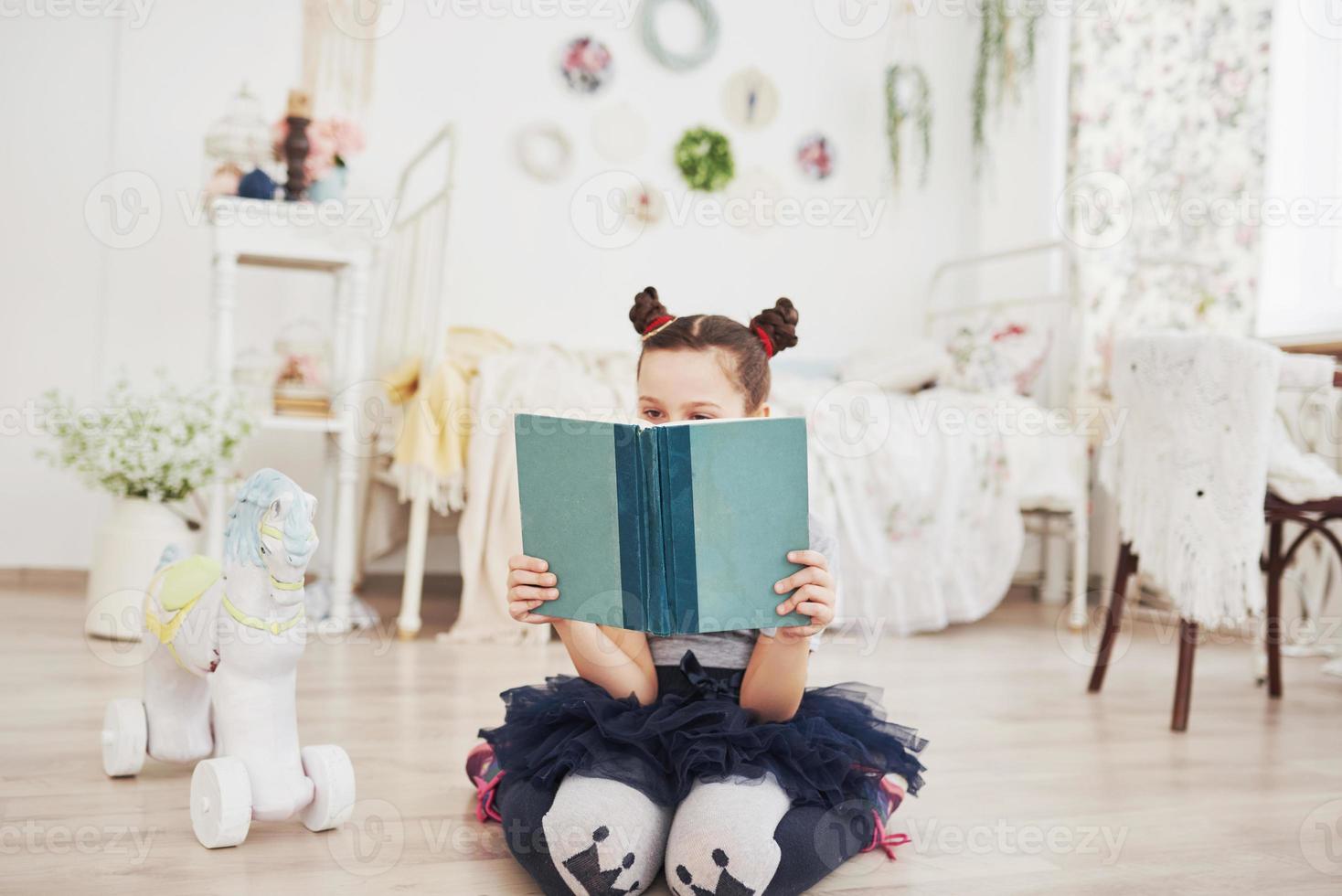 Cute little child girl reading a book in the bedroom. Kid with crown sitting on the bed near window photo