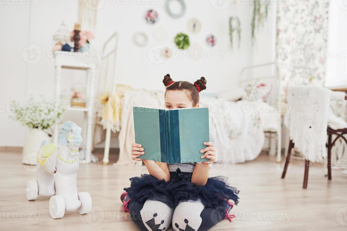 niña linda niña leyendo un libro en el dormitorio. niño con corona sentado en la cama cerca de la ventana foto