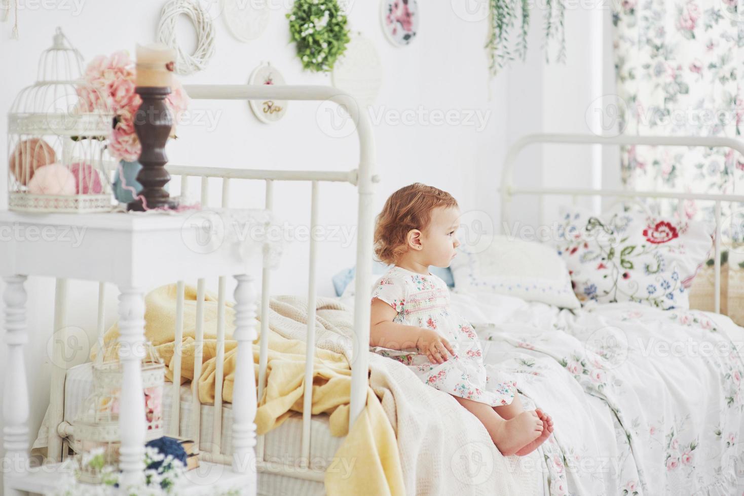 concepto de infancia. niña en vestido lindo emplazamiento en la cama jugando con juguetes en la casa. habitación infantil blanca vintage foto