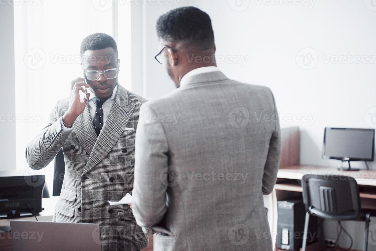 empresario moderno en el trabajo. Dos hombres de negocios confiados en ropa formal discutiendo algo mientras uno de ellos mira los documentos y el segundo una importante llamada telefónica. foto