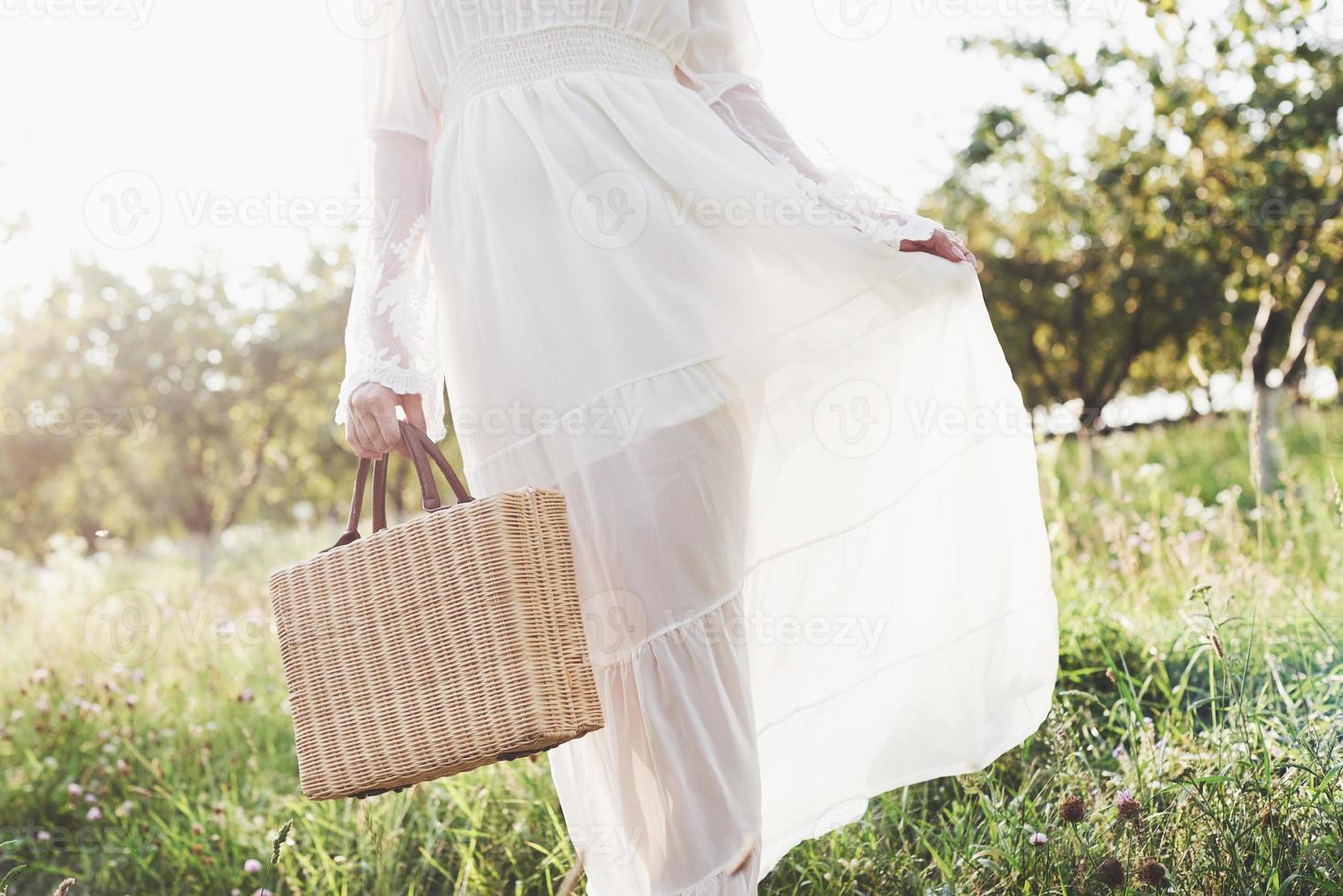 A young girl in a white long dress is walking in the garden. Beautiful sunset through the leaves of trees photo