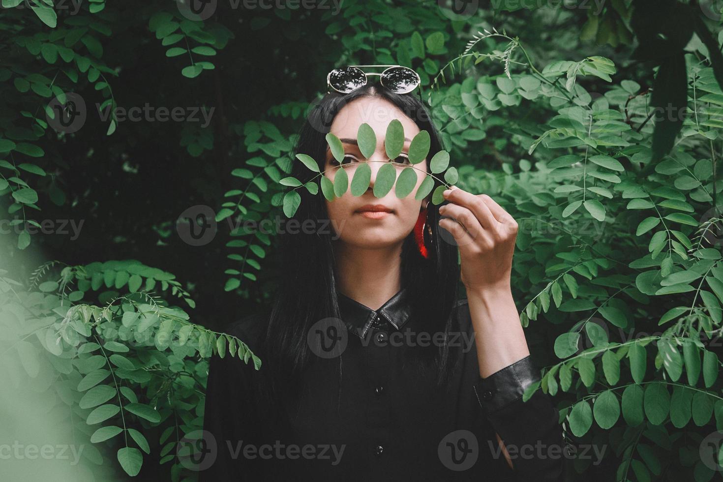 Woman in black clothes and glasses holds a twig in her hand on background of green leaves photo