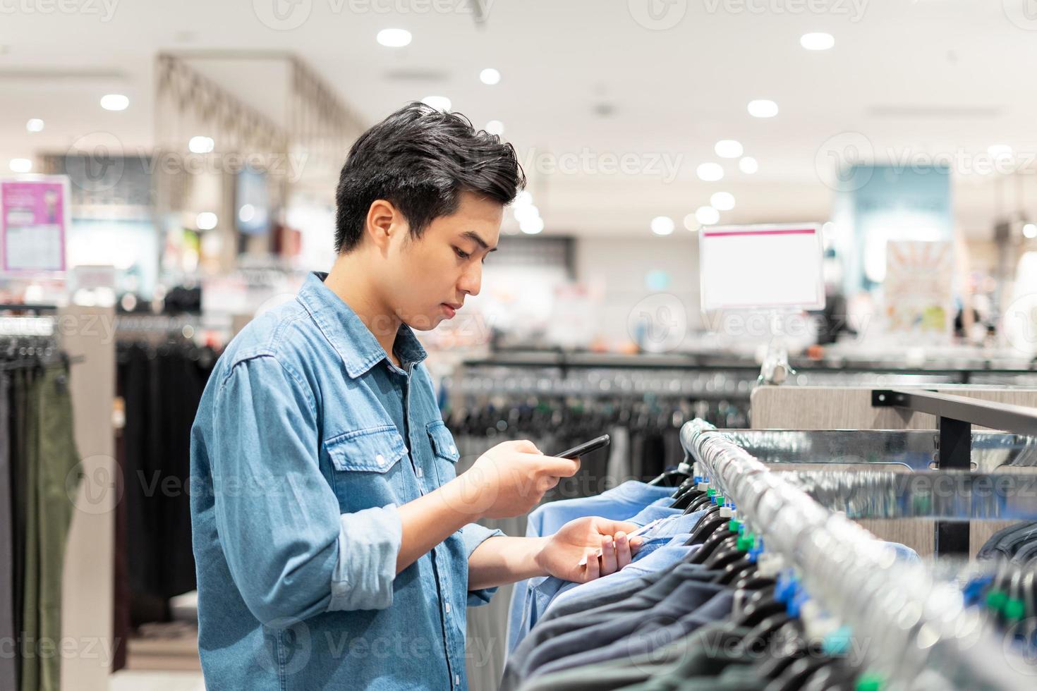 Hombre inteligente asiático que usa su teléfono está recogiendo ropa en la tienda de ropa en el centro comercial, foto