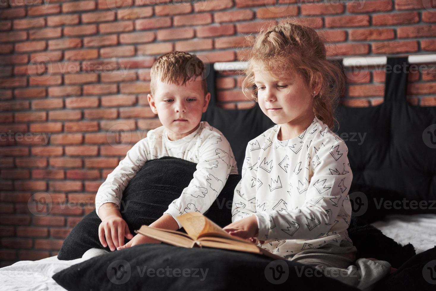 dos niños se acuestan en una cama grande y leen un libro interesante. están vestidos con el mismo pijama foto