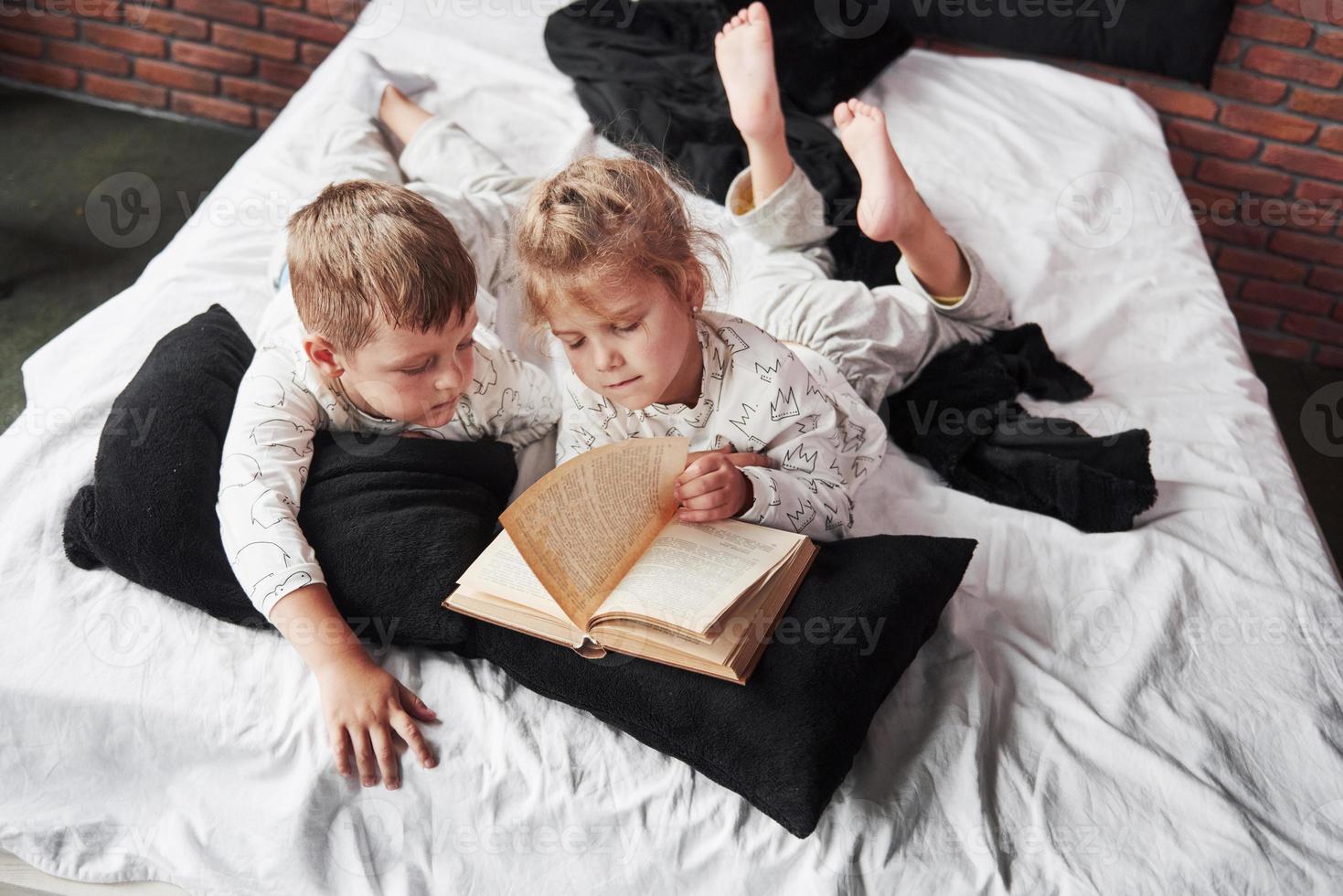 dos niños se acuestan en una cama grande y leen un libro interesante. están vestidos con el mismo pijama foto