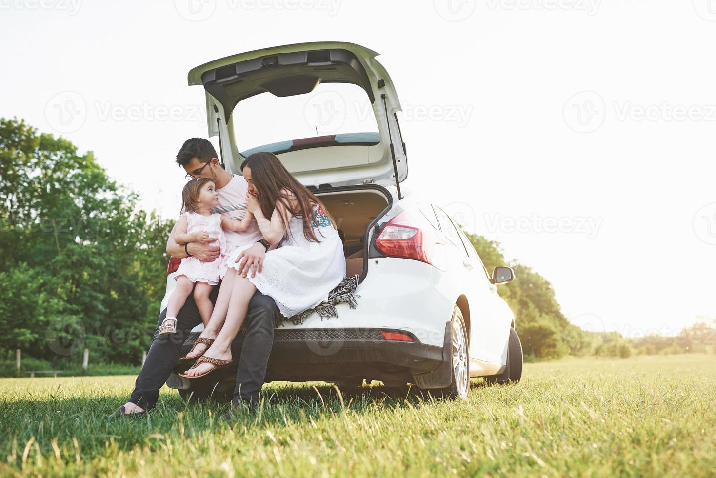 Pretty young married couple and their daughter are resting in the nature. The mother father and little girl are sitting on open car boot photo