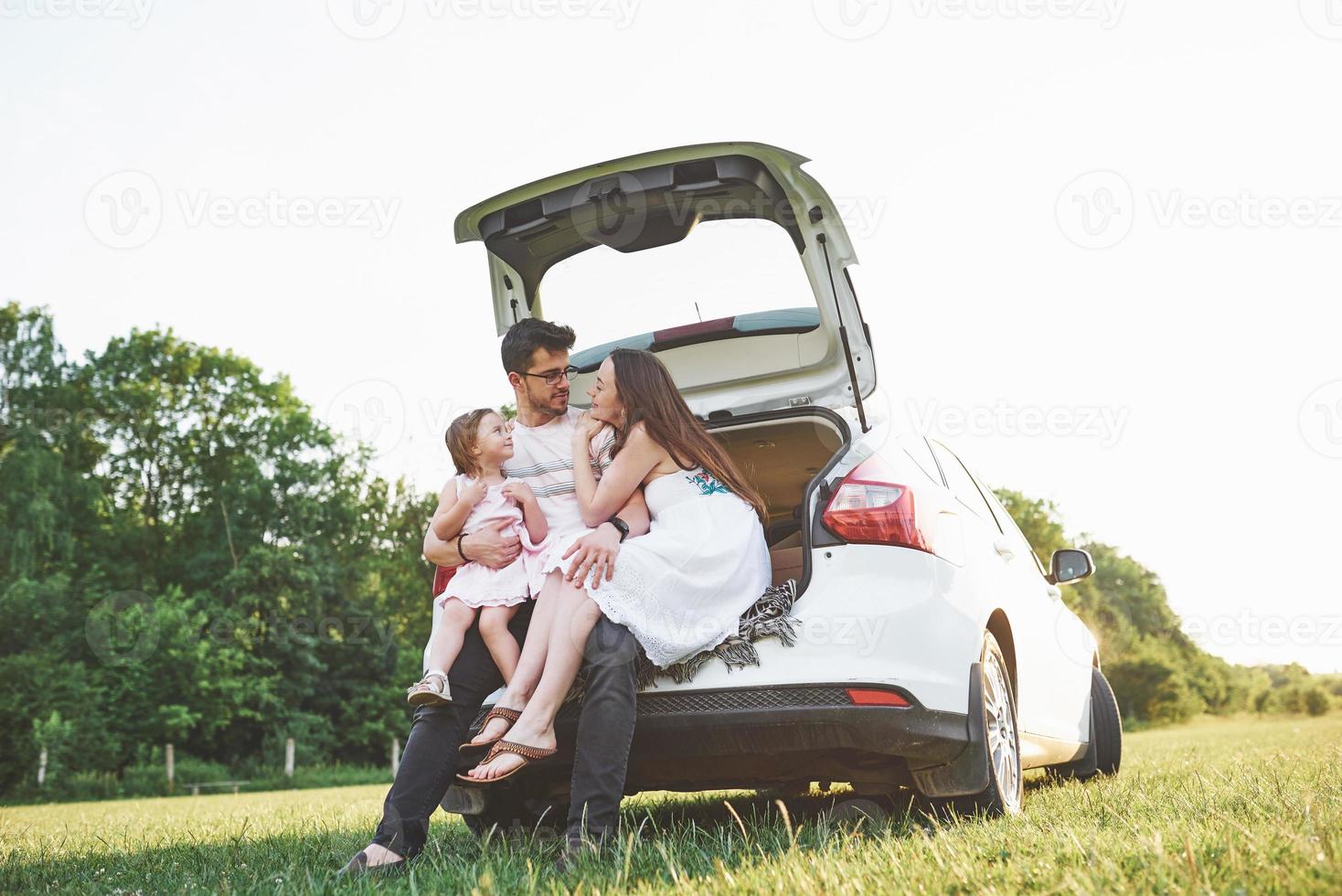 bastante joven matrimonio y su hija descansan en la naturaleza. la madre, el padre y la niña están sentados en el maletero del coche abierto foto