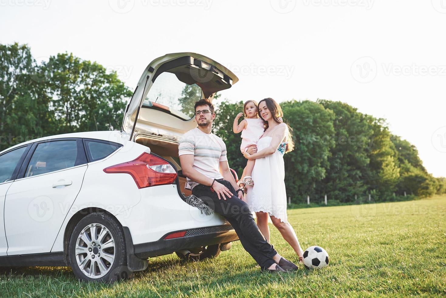 bastante joven matrimonio y su hija descansan en la naturaleza. la madre, el padre y la niña están sentados en el maletero del coche abierto foto