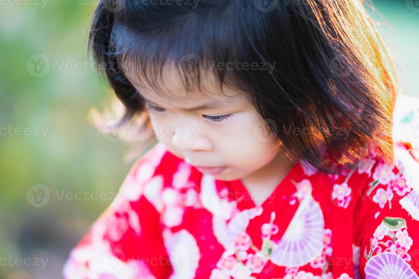 Close up, adorable 1-2 year old Asian girl is looking down at her path. A cheerful baby wears a red Japanese-style dress. During the summer or spring. photo