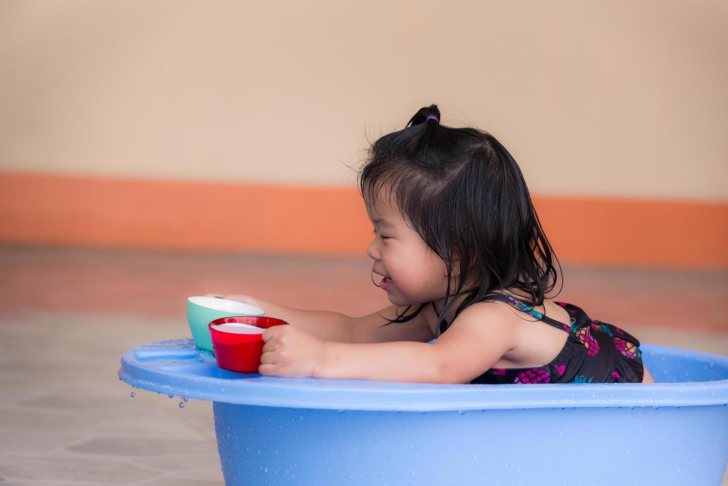 Baby wearing black swimsuit is playing in blue basin. Girl played two glasses that were used to scoop water and to transfer it back and forth. Child smile and have fun with wet body. Kids 2-3 year old photo