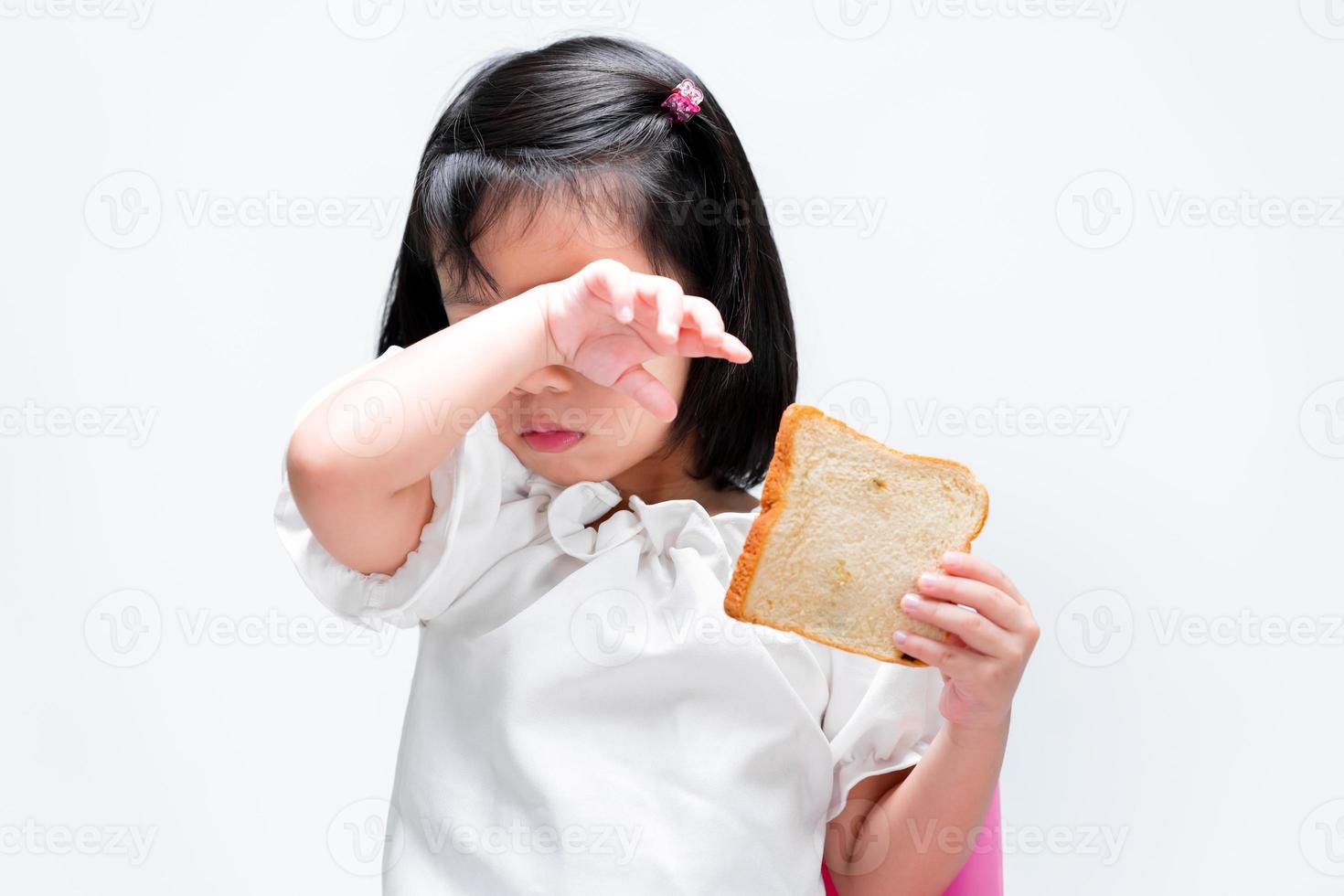 A cute little girl rubs her eyes from the back of her wrists due to itching or irritation. Child holding bread. Food concepts and food allergies. photo