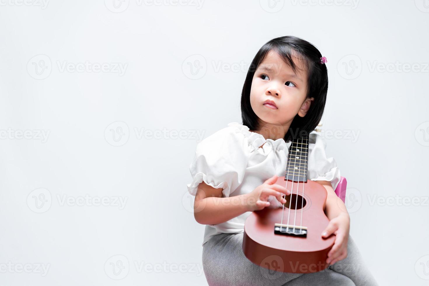 Adorable child girl puts her ukulele on her shoulder. Kid play musical instruments, looking sideways. On studio white background. photo