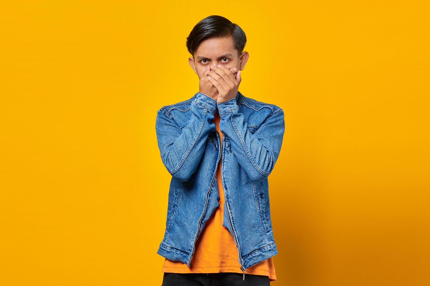 Portrait of surprised young Asian man covering mouth with hands over yellow background photo