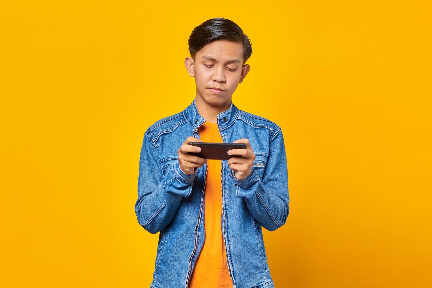 Hombre asiático con teléfono móvil jugando con cara enojada sobre fondo amarillo foto