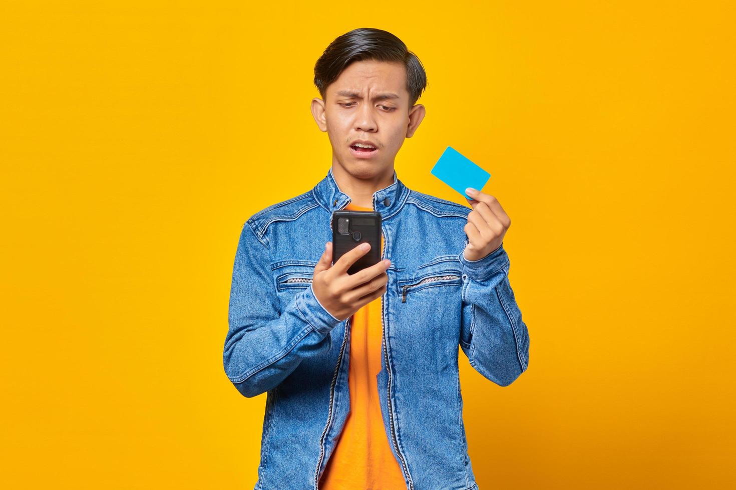 Shocked asian man holding mobile phone and showing credit card over yellow background photo