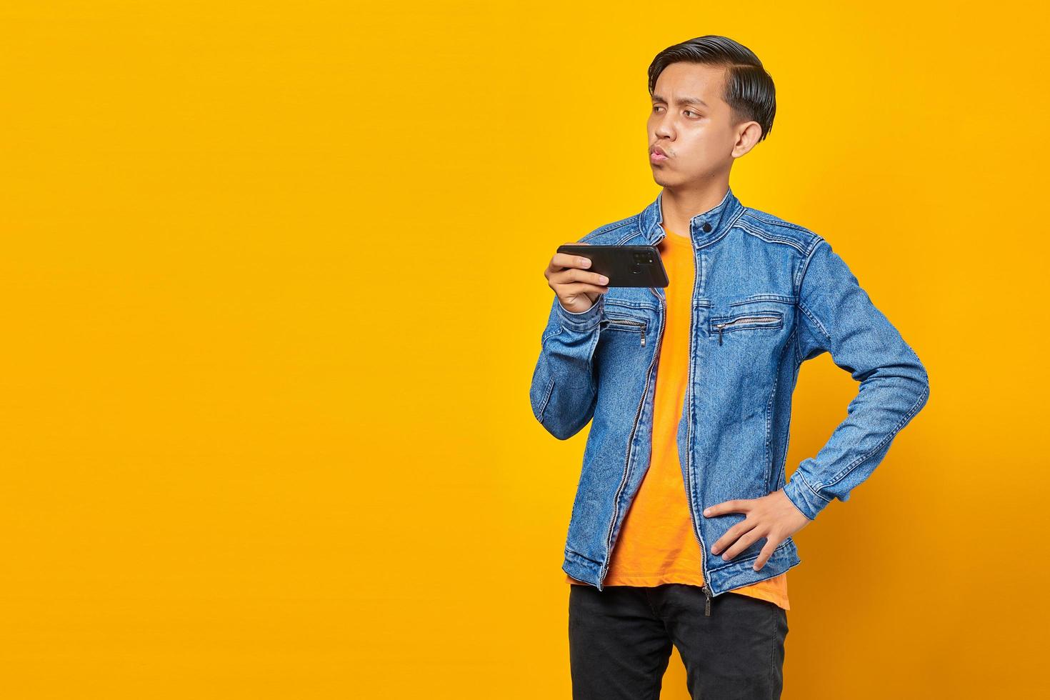 Hombre asiático con teléfono móvil jugando con cara enojada sobre fondo amarillo foto