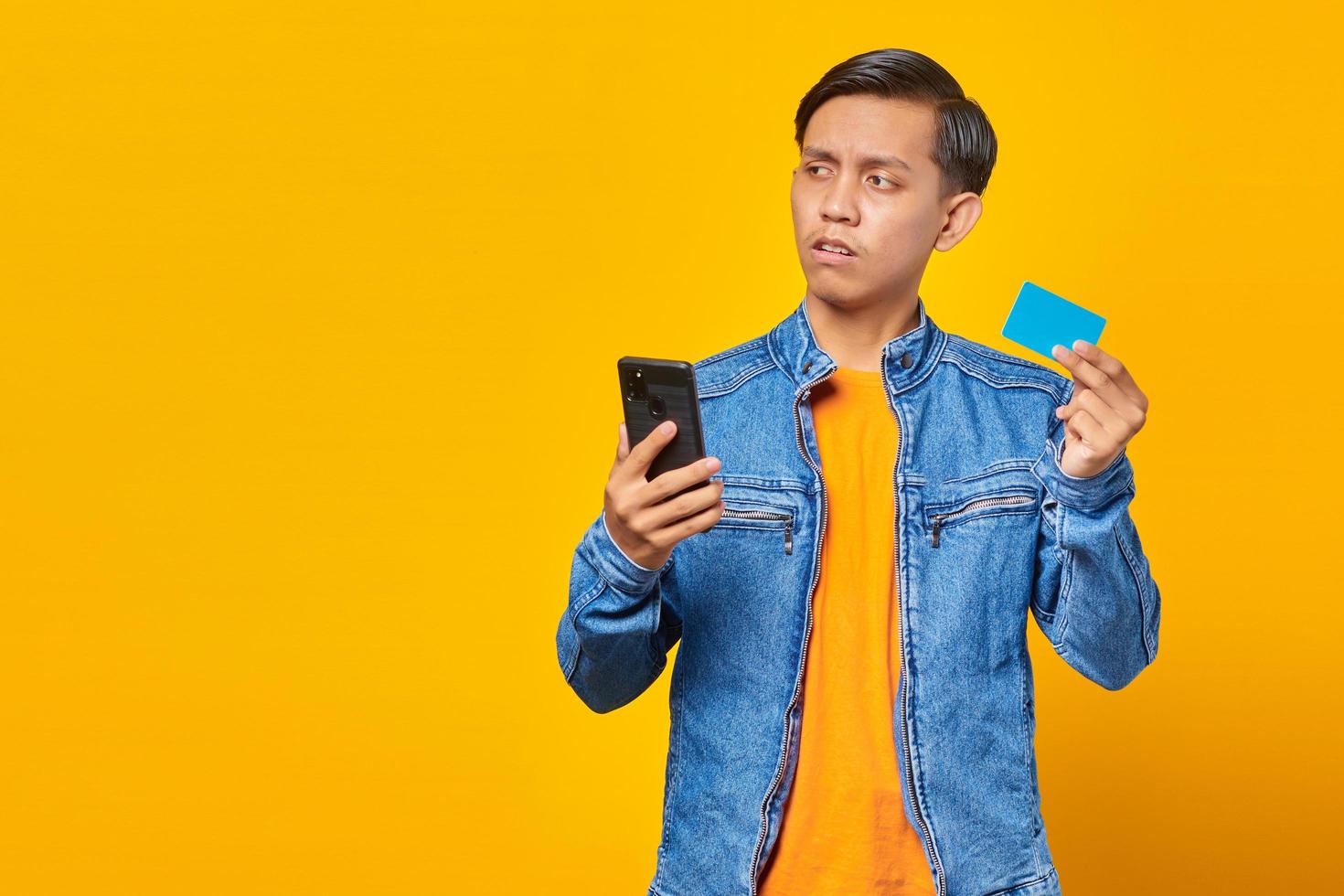 Conmocionado hombre asiático sosteniendo teléfono móvil y mostrando tarjeta de crédito sobre fondo amarillo foto