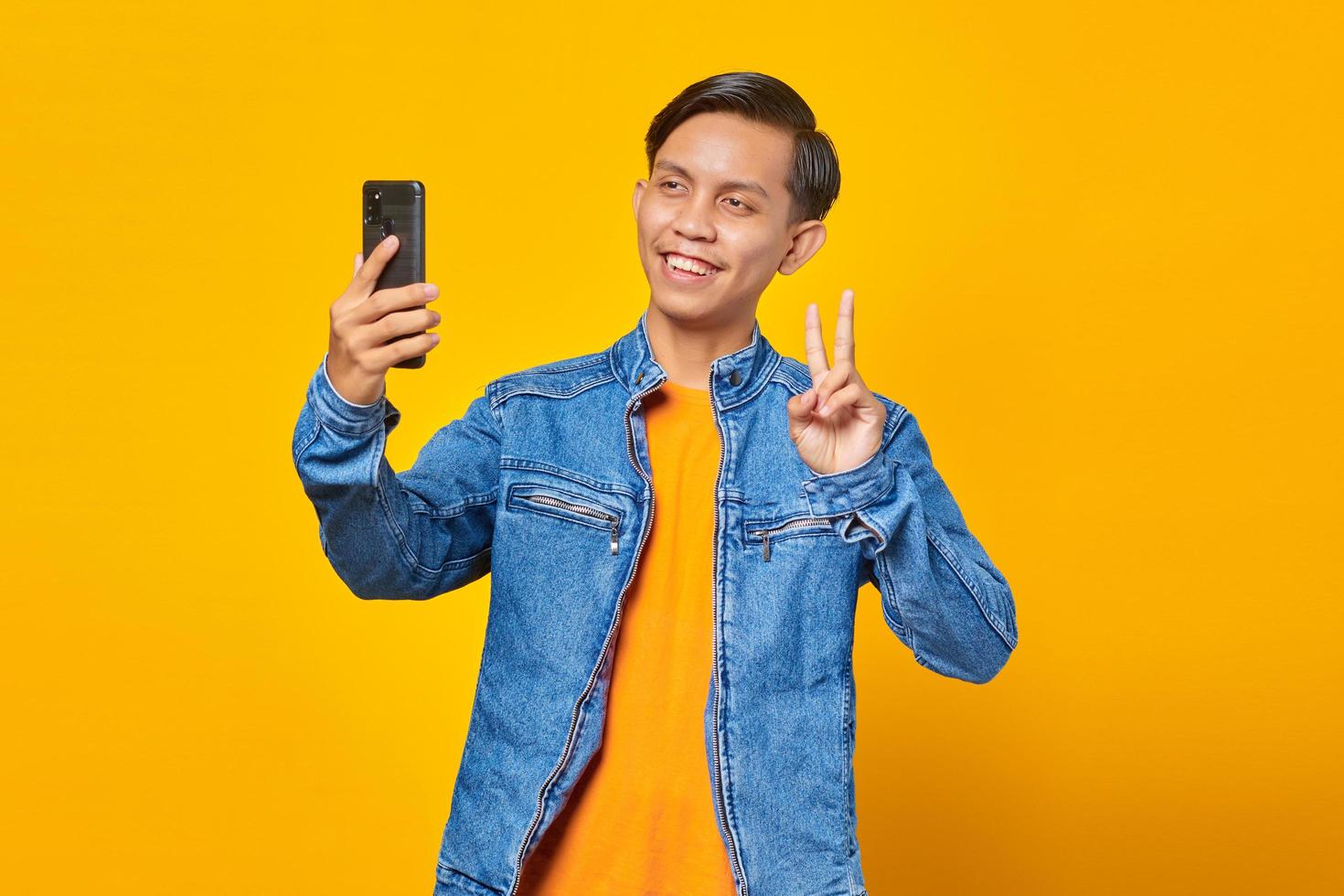 portrait of asian man smiling and showing peace sign while taking selfie on mobile isolated on yellow background photo