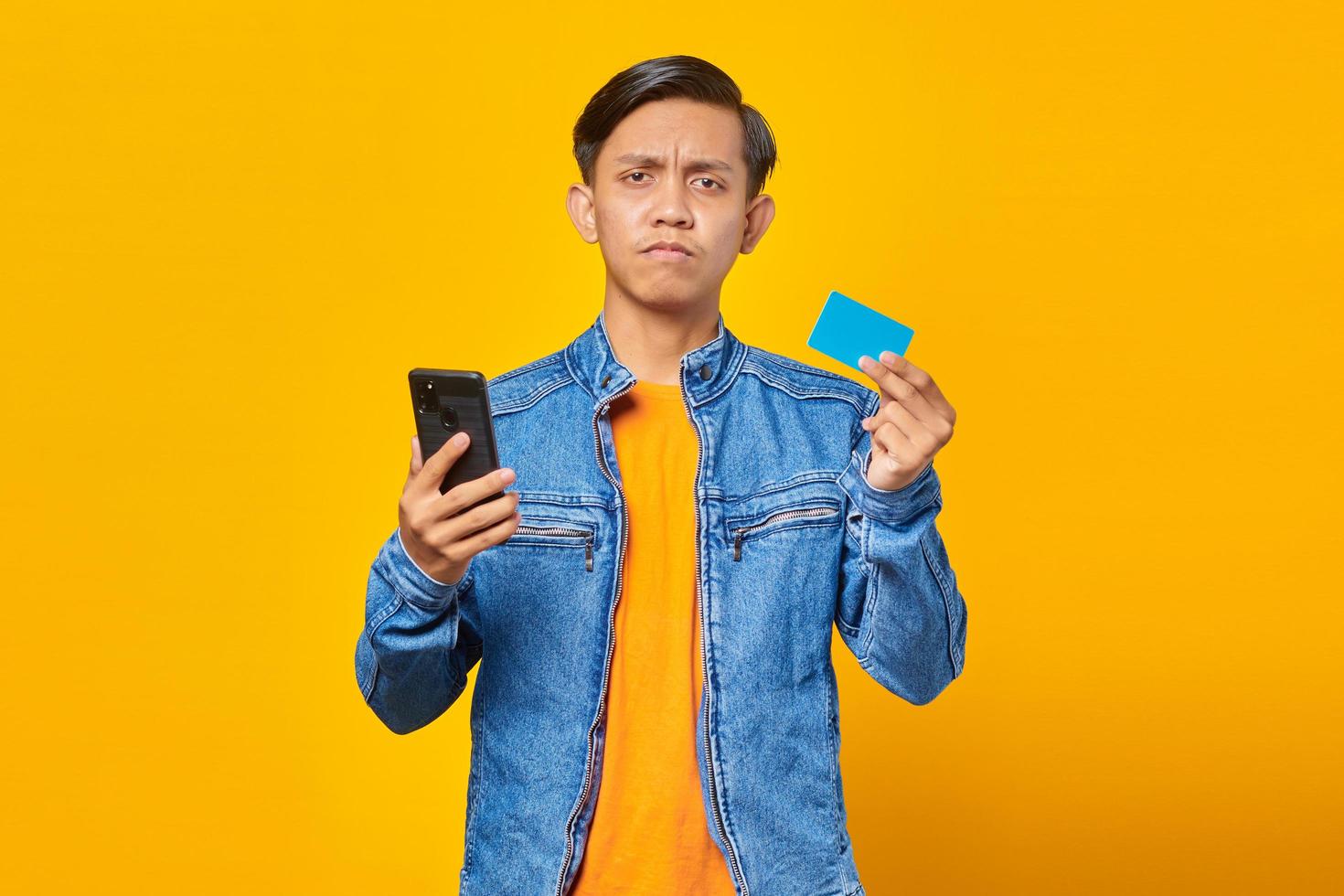 Shocked asian man holding mobile phone and showing credit card over yellow background photo