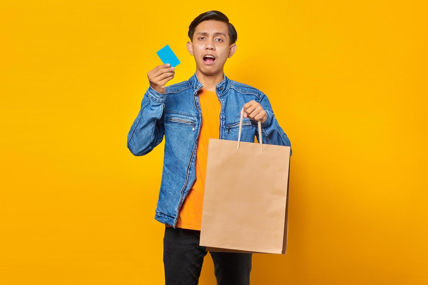 Portrait of shocked asian man holding credit card and shopping bag over yellow background photo